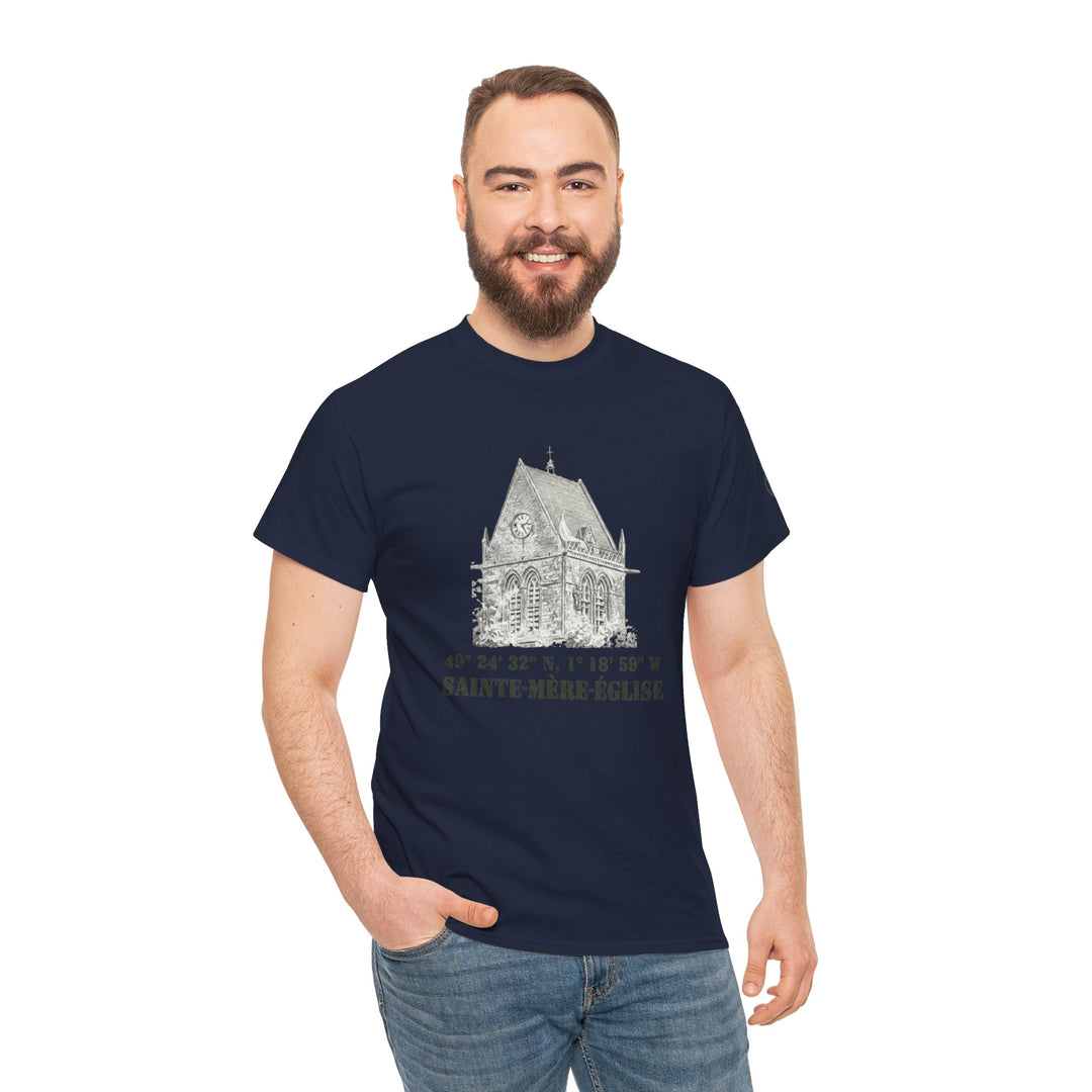 Navy t-shirt by Veteran owned, featuring a church graphic with coordinates and "SAINTE-MÈRE-ÉGLISE" lettering. Worn with denim jeans by a smiling man.