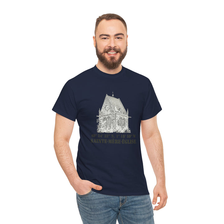 Navy t-shirt by Veteran owned, featuring a church graphic with coordinates and "SAINTE-MÈRE-ÉGLISE" lettering. Worn with denim jeans by a smiling man.